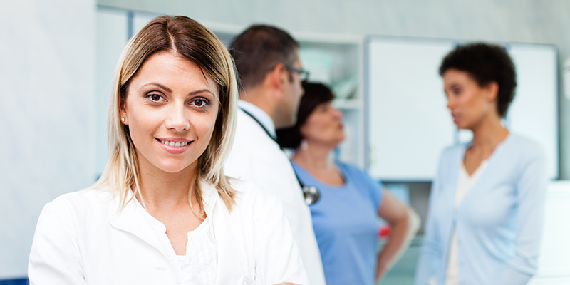 Image of woman in medical setting smiling at the camera, group of medical staff and a patient in conversation in the background.
