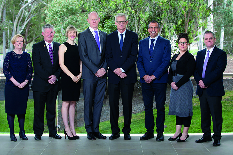 All members of the Executive Group standing infront of a glass panel railing with a leafy backdrop