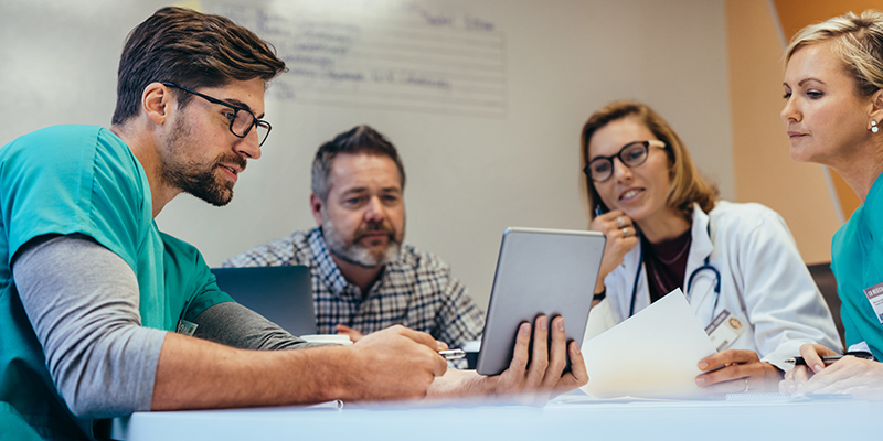 Group of medical professionals sitting arounf a desk all focussed on a tablet being held by one of the professionals