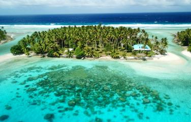 Landscape photo of a small tropical island in the Marshall Islands covered in lush green palm trees, surrounded by white sand and clear pale blue-green water. In the background there is a strip of dark blue ocean leading to the horizon and cloudy blue sky