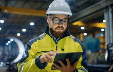 An engineer wearing a hard hat and high visibility jacket is looking at a tablet