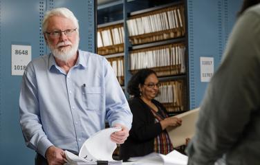 Man cycling through papers while looking up to a person out of focus with many files and books on a shelf in the background.