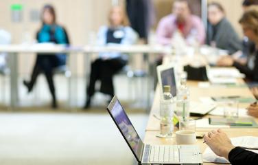 A room full of people out of focus sitting around desks, with a close up of a laptop.