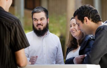 A male and female smiling and presenting in a cheerful manner engaged in conversation with two other males across a table in an outdoor setting.
