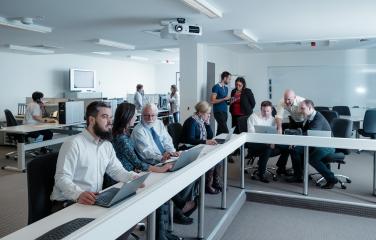 An office showing 13 people sitting in an open plan area working on laptops in pairs and small groups. In the background two of the people are sitting at desks taking phone calls.