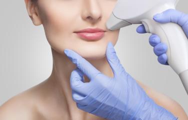 A woman is undergoing hair removal treatment on her upper lip by a gloved service provider with a device.