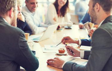 Several people dressed in business attire having a conversation around a wooden table.