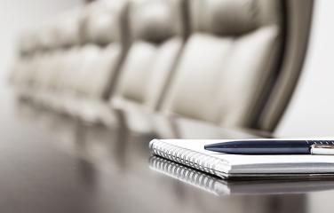 A pen and notepad sitting atop a wooden table with blurred out chairs in the distance.