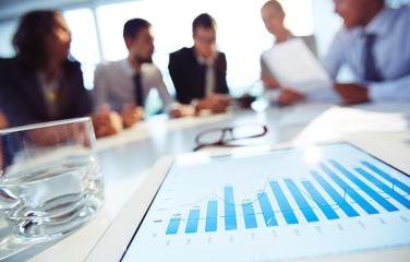 Five people in business attire having a meeting out of focus with a close-up of a tablet displaying a graph.