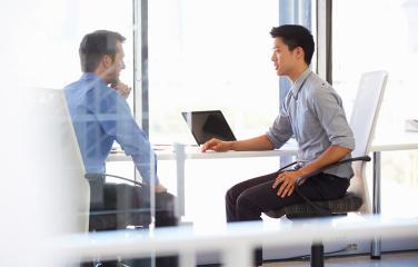 Two middle aged men sitting at a desk having a discrete conversation.