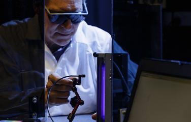 A male scientist wearing a white lab coat and dark eye protecting glasses leans forward to adjust an ultraviolet radiation measuring device which is positioned to measure the ultraviolet radiation coming from a black light.