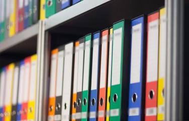 Many different coloured folders sitting on a bookshelf.