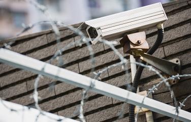 A closed circuit camera behind a fence topped with barbed wire.