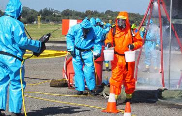 A person wearing a full body protection suit holding two buckets carrying out a simulated emergency response with others in protective clothing in the background assisting and looking on.