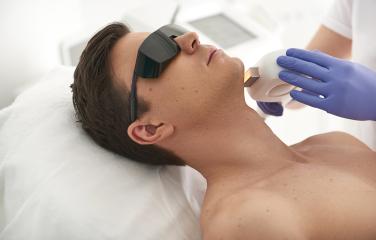 Man lying on medical bed, wearing dark black eye glasses while receiving laser treatment on his chin from a practitioner wearing blue latex gloves