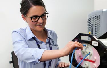 A happy confident female scientist leans forward to adjust a cable with her right hand which leads to a phantom used in calibrations of radiotherapy equipment.