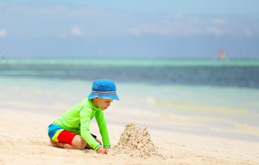 A young boy playing with sand on a beach wearing a wide-brim hat, long-sleeve shirt and shorts.