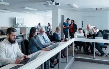 Several people gathered around a long desk discussing with each other in small groups around laptops. Two people are focussing on their laptops working individually.