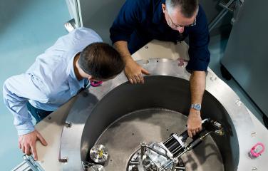 Alt text – From above, a male inspector and a male scientist peer into a metal cylinder with a diameter of two meters. The male scientist reaches down to adjust the scientific equipment at the bottom of the cylinder.