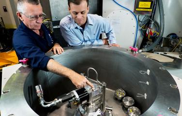 A male inspector and a male scientist peer into a metal cylinder with a diameter of two meters. The male scientist points at the scientific equipment at the bottom of the cylinder.