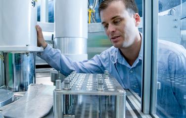 A male inspector leans forward to get a better look at scientific equipment that is shielded by transparent plastic. He reaches out with his right hand to balance himself on a metal cylinder.