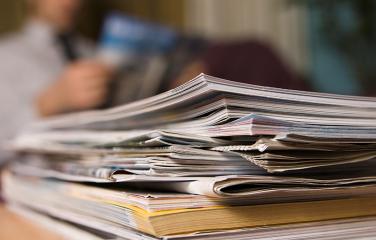 A stack of publications on a wooden table with an out of focus man reading in the background.