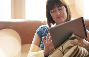 Middle-aged woman sitting on a couch looking insightful while browsing on a tablet.