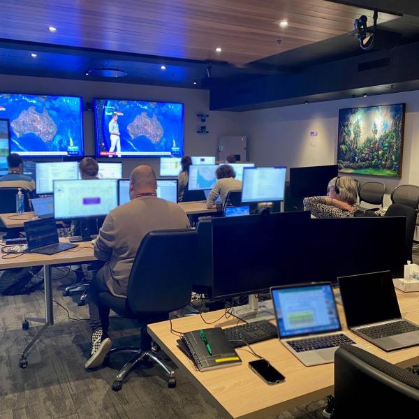 A group of people are sitting in a room looking at computer monitors and TV screens