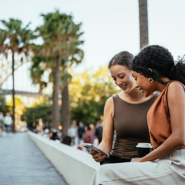 Two women enjoying a sunny day out near Sydney Harbour Bridge, smiling and talking as they look at the smart phone