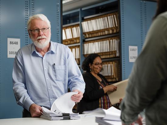 Man cycling through papers while looking up to a person out of focus with many files and books on a shelf in the background.