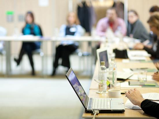 A room full of people out of focus sitting around desks, with a close up of a laptop.