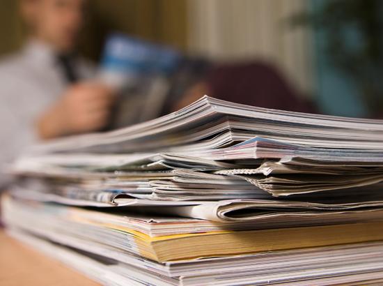 A stack of publications on a wooden table with  an out of focus man reading in the background.
