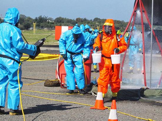 A person wearing a full body protection suit holding two buckets carrying out a simulated emergency response with others in protective clothing in the background assisting and looking on.